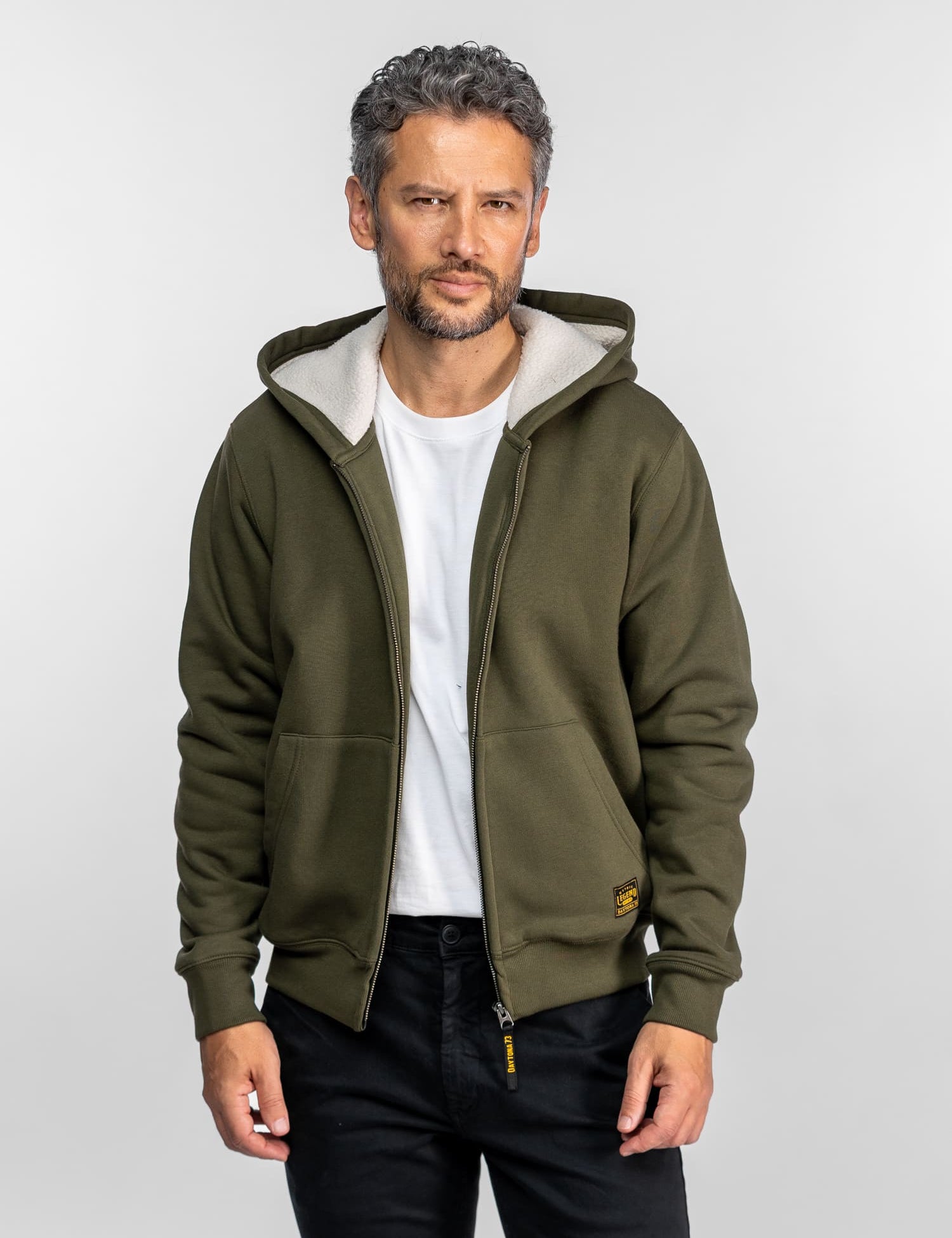 Un homme vêtu d'un sweat à capuche vert olive et d'une chemise blanche se tient avec assurance sur un fond uni, les mains dans les poches, regardant l'objectif.