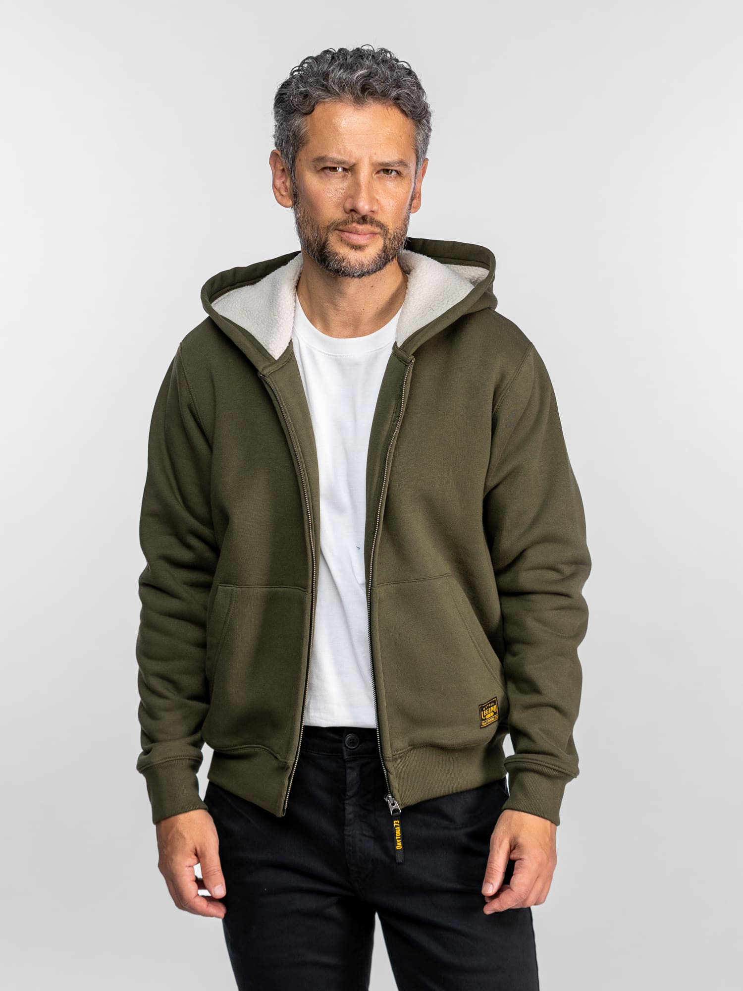 Un homme vêtu d'un sweat à capuche vert olive et d'une chemise blanche se tient avec assurance sur un fond uni, les mains dans les poches, regardant l'objectif.
