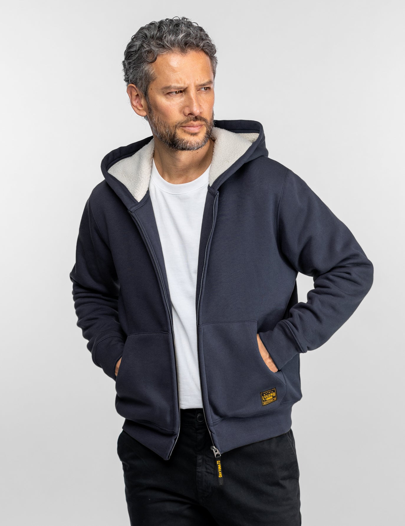 Homme en sweat-shirt bleu marine sur chemise blanche, regardant sur le côté. Mains dans les poches, pose décontractée, fond neutre.