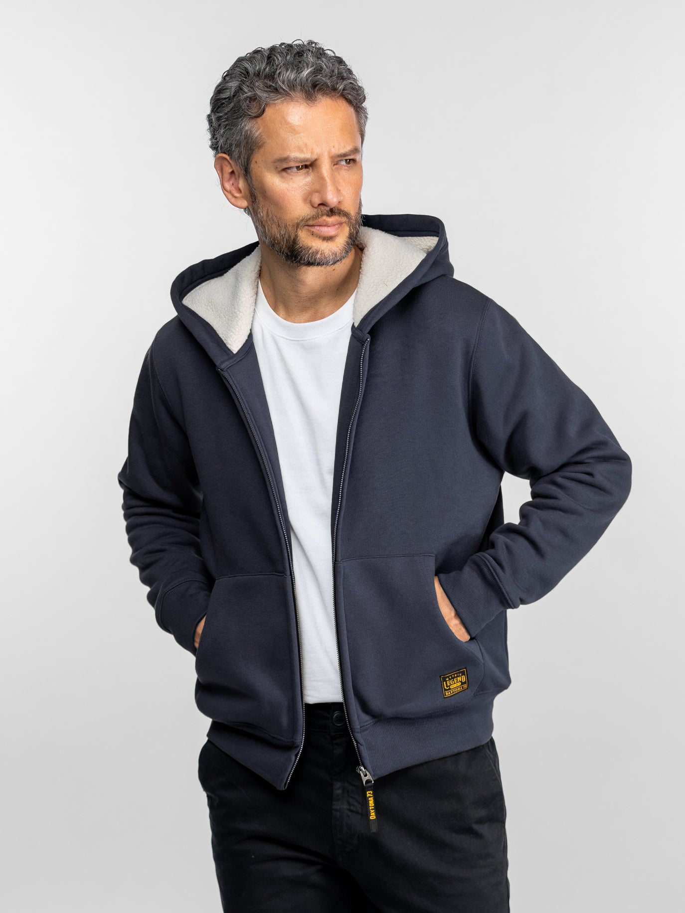 Homme en sweat-shirt bleu marine sur chemise blanche, regardant sur le côté. Mains dans les poches, pose décontractée, fond neutre.