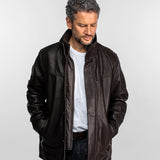 Un homme vêtu d'une veste en cuir marron et d'une chemise blanche se tient devant un fond uni, les mains dans les poches, le regard pensif.