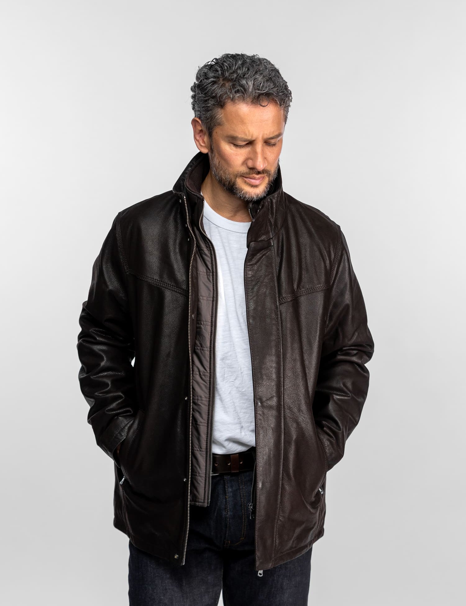 Un homme vêtu d'une veste en cuir marron et d'une chemise blanche se tient devant un fond uni, les mains dans les poches, le regard pensif.