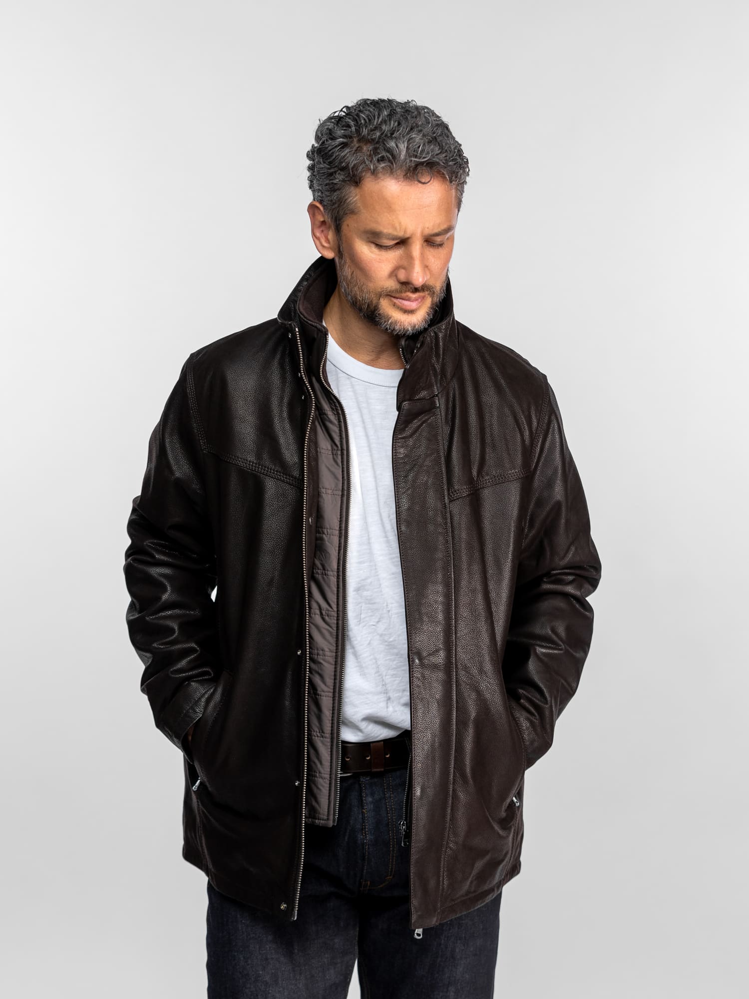 Un homme vêtu d'une veste en cuir marron et d'une chemise blanche se tient devant un fond uni, les mains dans les poches, le regard pensif.