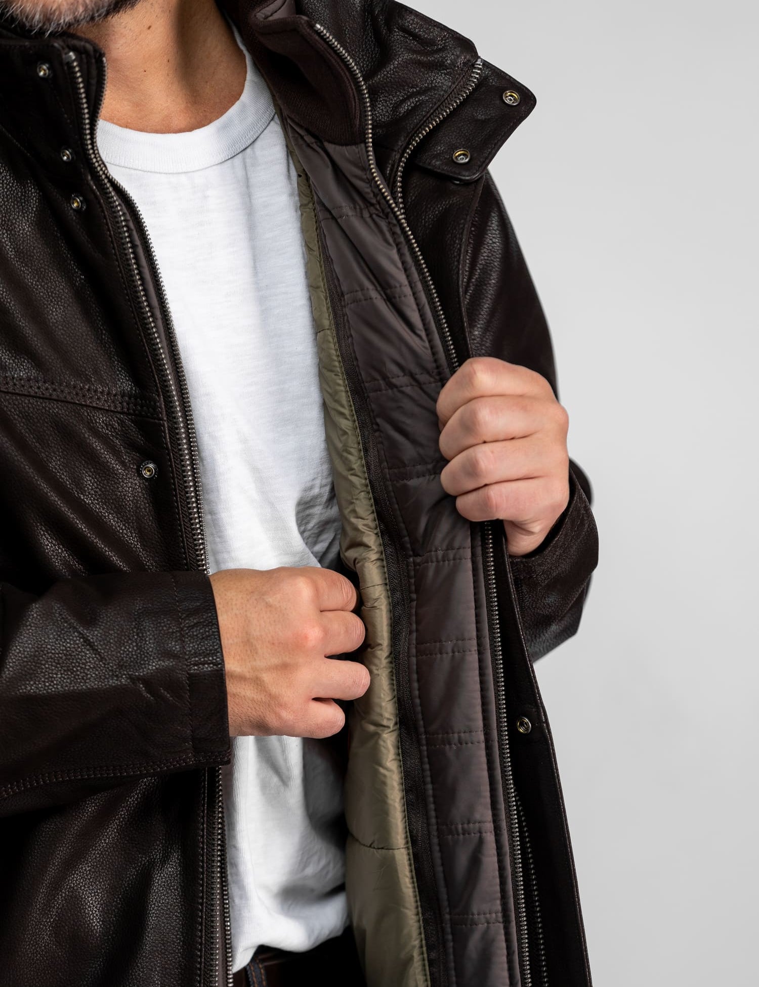 Un homme en chemise blanche ouvre la fermeture éclair d'une veste en cuir marron foncé, révélant une doublure intérieure matelassée, sur un fond neutre.