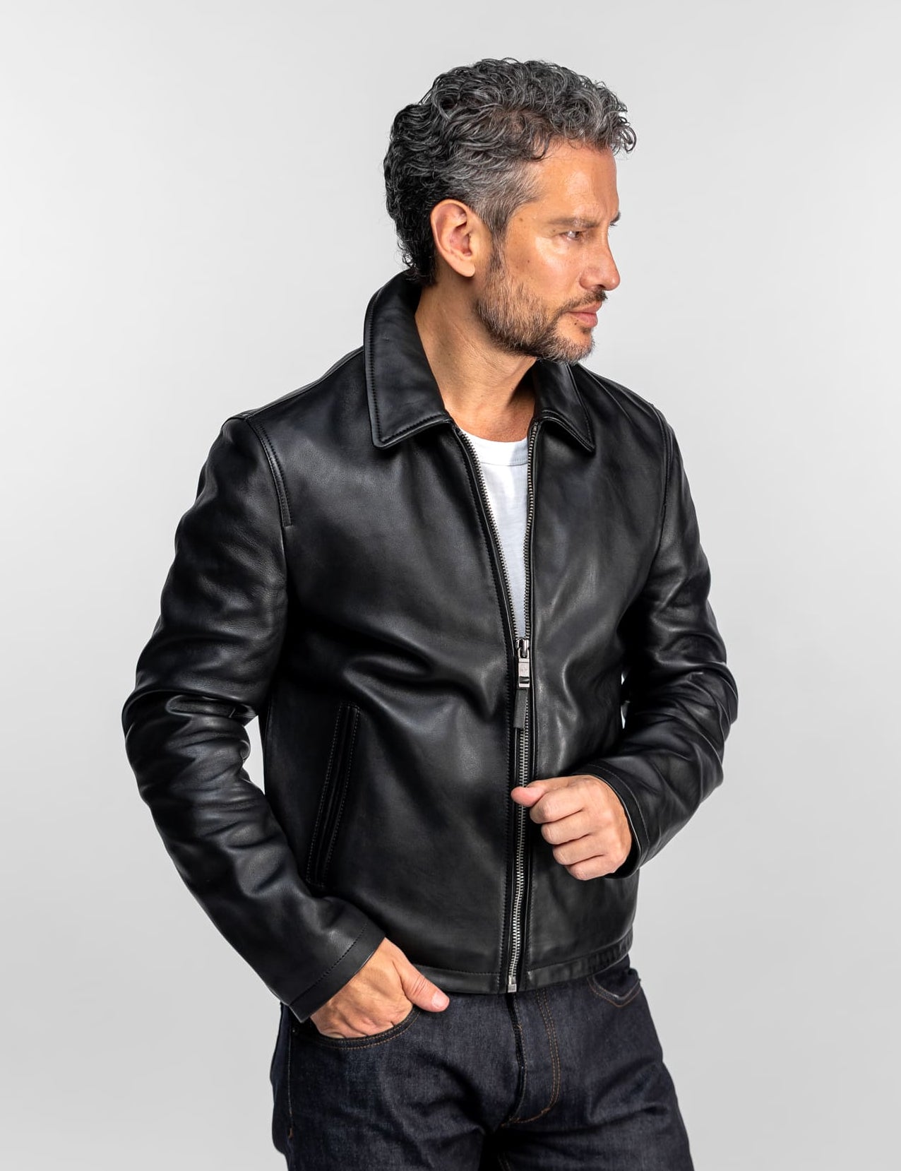 Un homme en veste de cuir noire et jean, regardant sur le côté sur un fond uni, dégageant un style décontracté et cool.