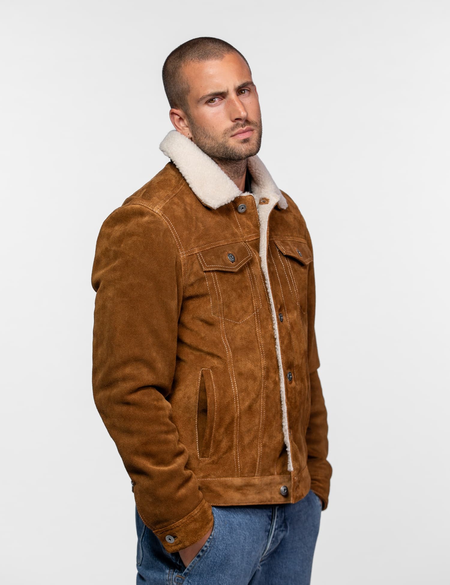 Man in a brown suede jacket with a shearling collar and blue jeans, standing against a plain background, looking serious.