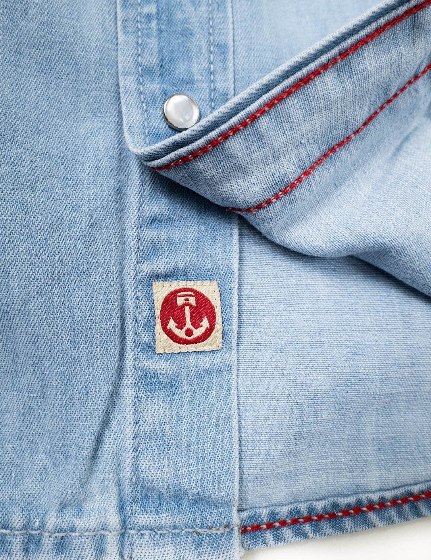 Close-up of a light blue denim shirt with red stitching and a small anchor logo patch near a silver button.
