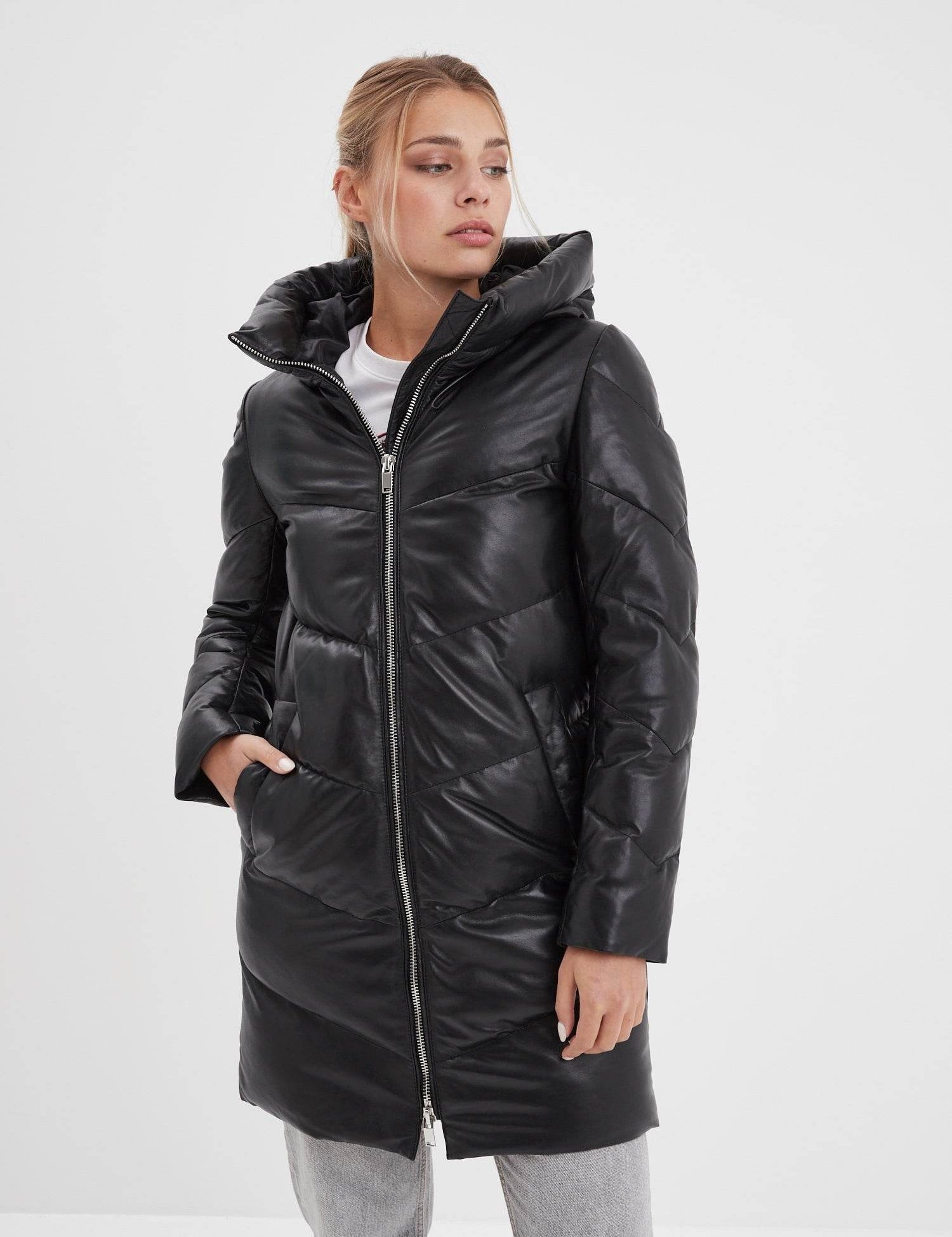 A woman in a black puffer coat with a hood stands against a white background, hands in pockets, looking to the side.