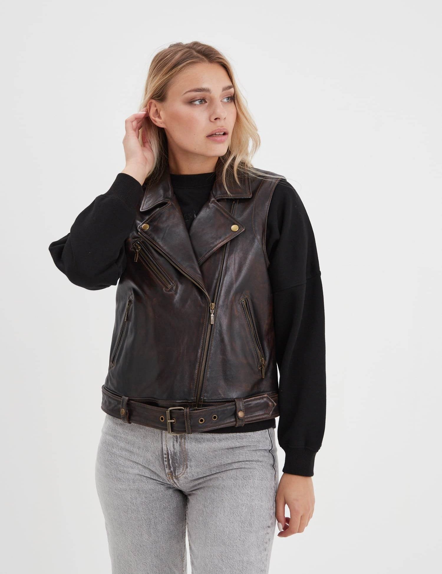 Woman in a black sweater and brown leather vest, looking to the side, with light jeans against a plain white background.