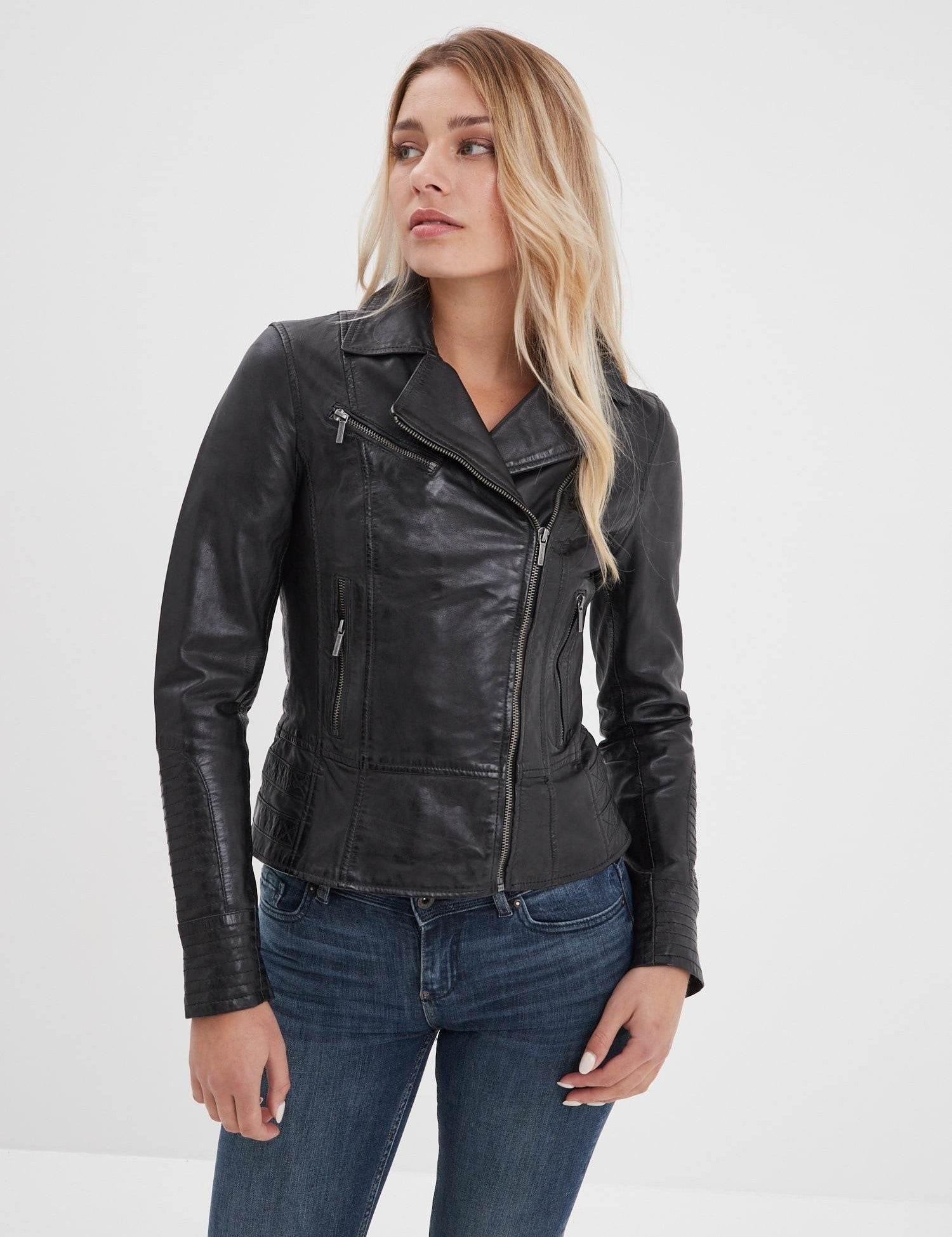 Woman in a black leather jacket and jeans gazes to the side against a plain white background, exuding a casual, stylish vibe.
