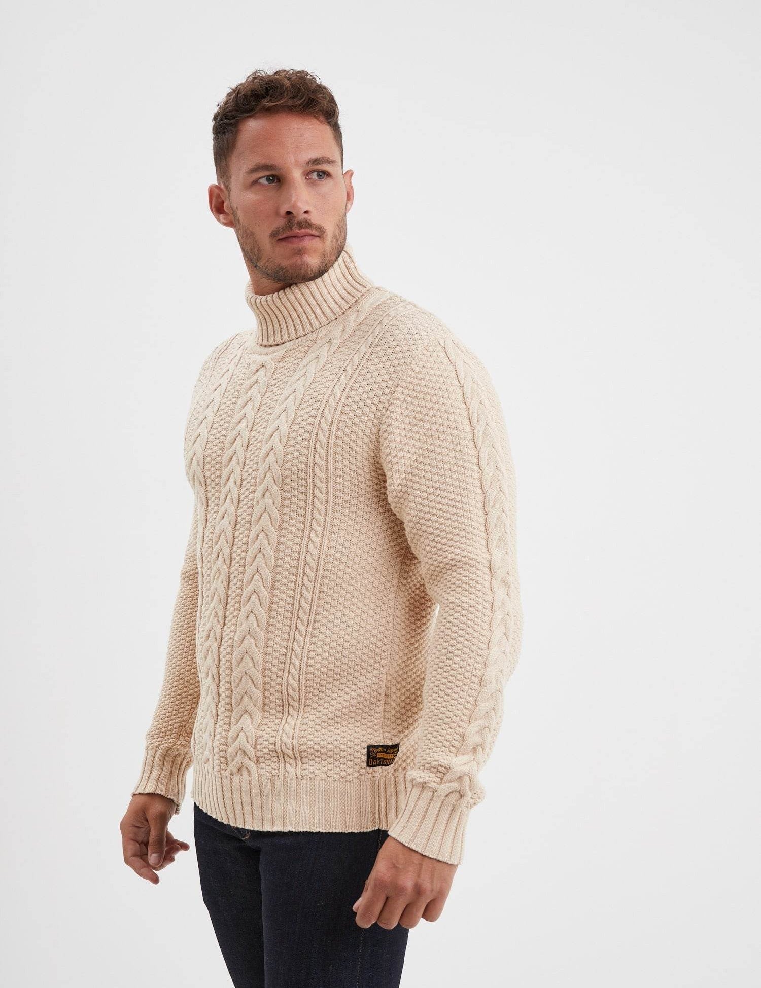 Pull beige à col roulé torsadé avec petit écusson noir.