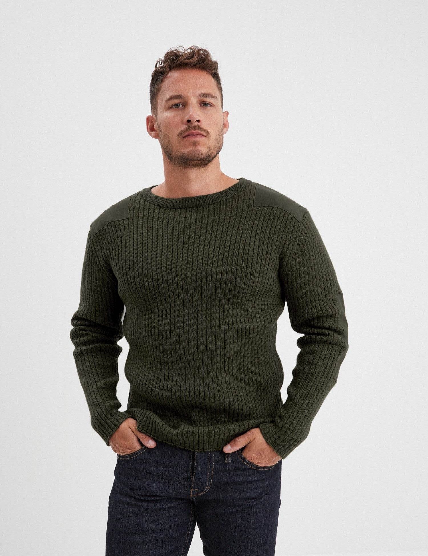 Man in a green ribbed sweater and jeans stands confidently against a plain background, hands in pockets, looking forward.