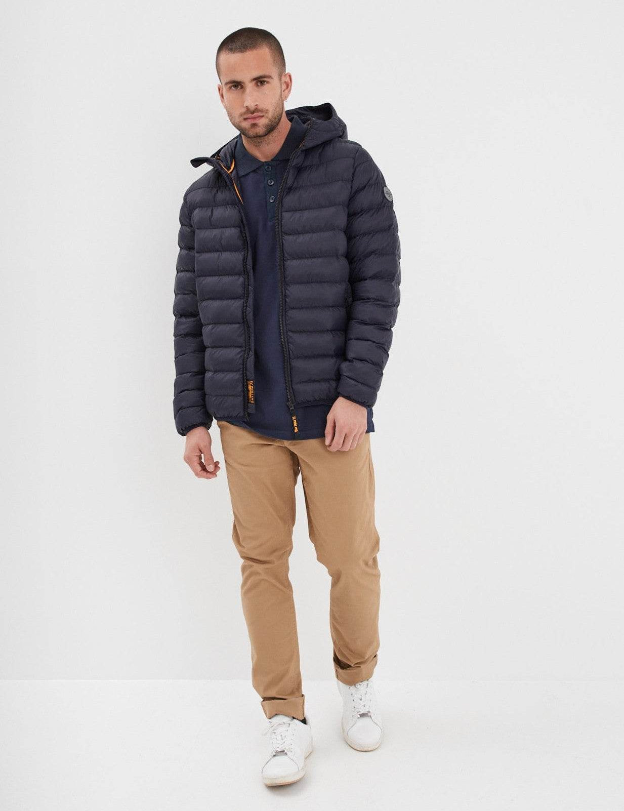 Man in a navy puffer jacket, beige pants, and white sneakers stands against a white background, looking at the camera.