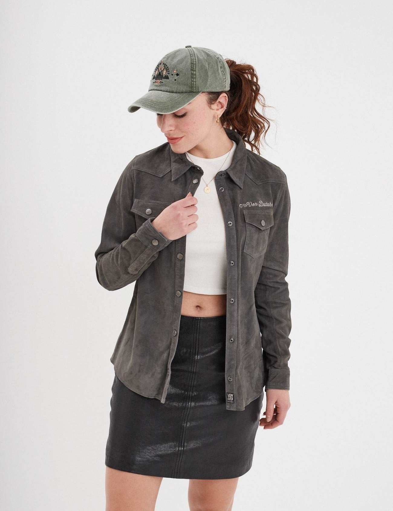 Woman in a green cap and gray jacket over a white top with a black skirt, looking down against a plain background.