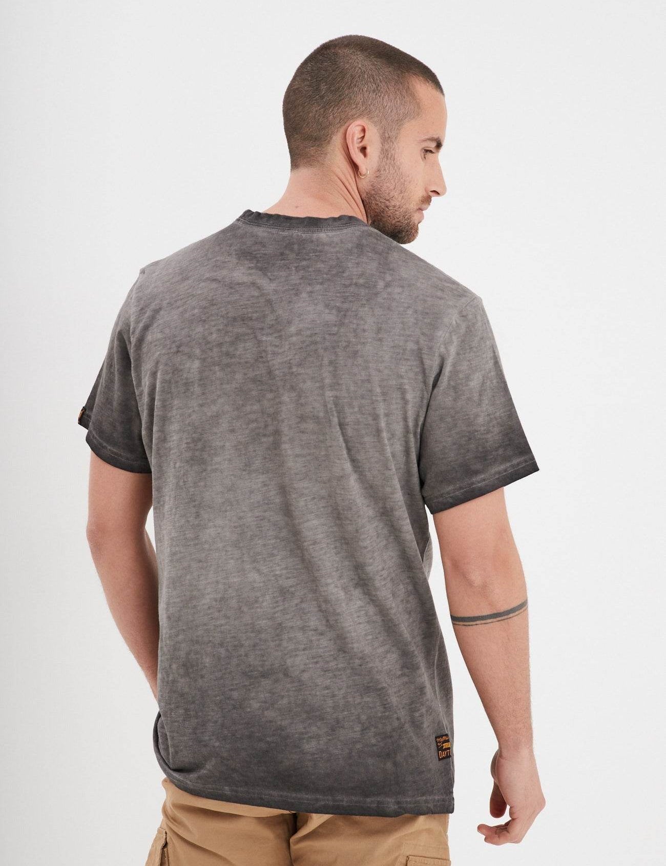 Man in a gray t-shirt and khaki shorts, facing away, with a tattooed arm, stands against a plain white background.