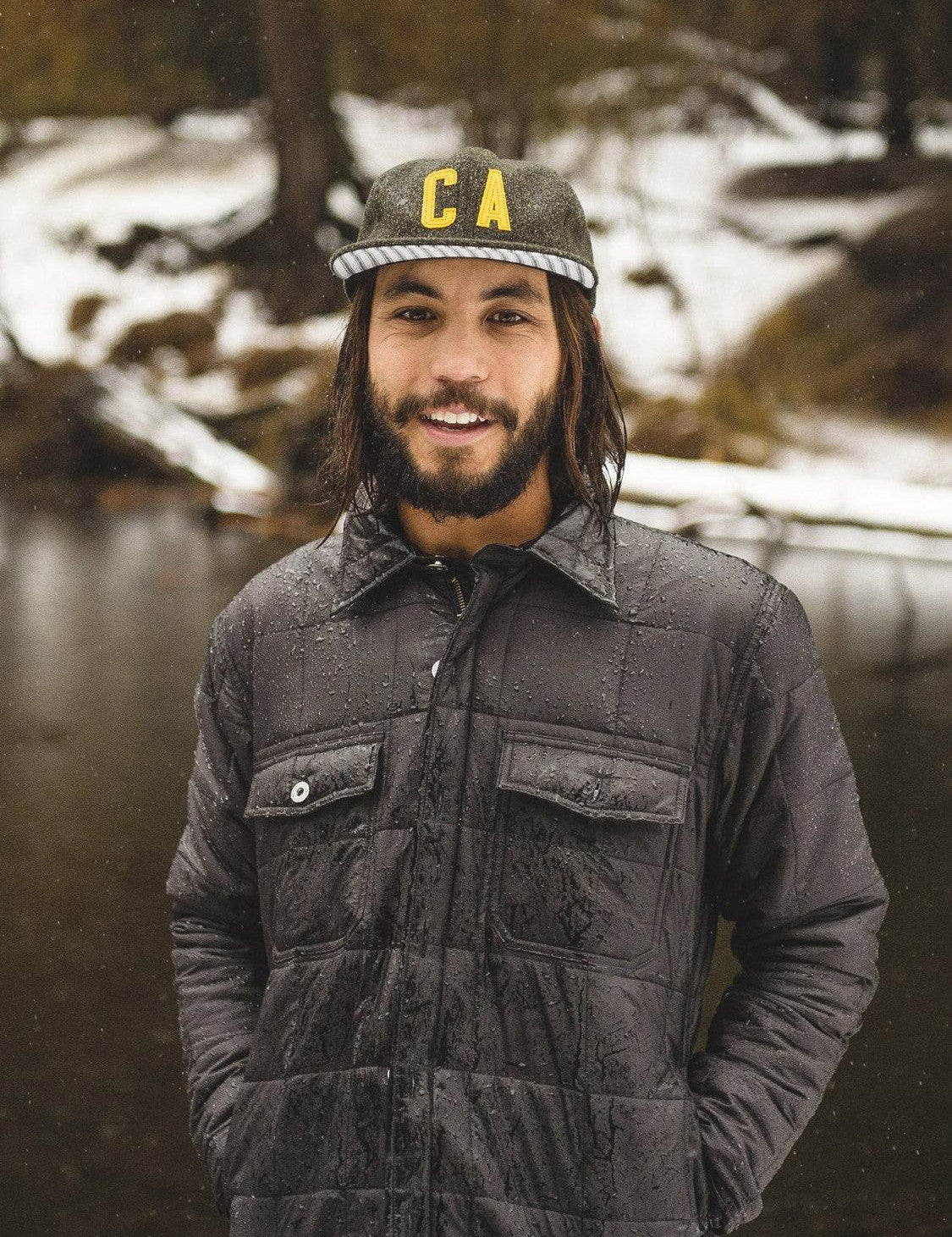 A man in a wet jacket and "CA" cap stands smiling by a snowy riverbank, blending nature and style in a serene outdoor setting.