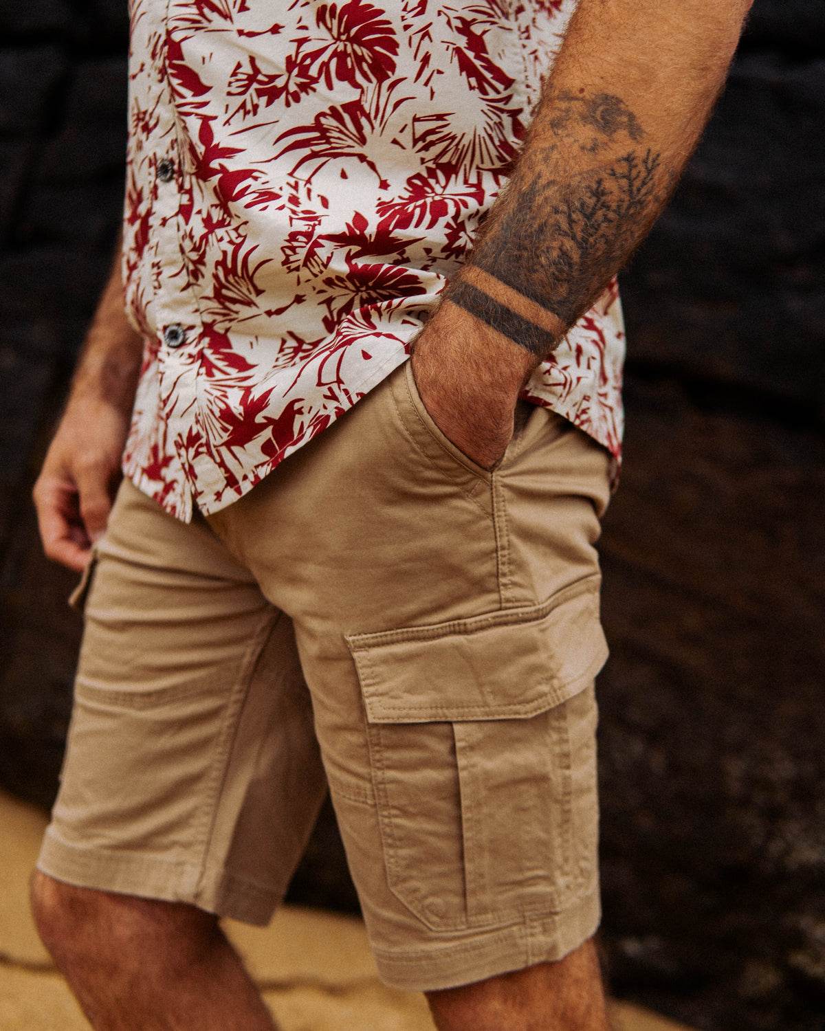 Man in floral shirt and cargo shorts with tattooed arm stands on sandy beach, hand in pocket, against dark rock background.