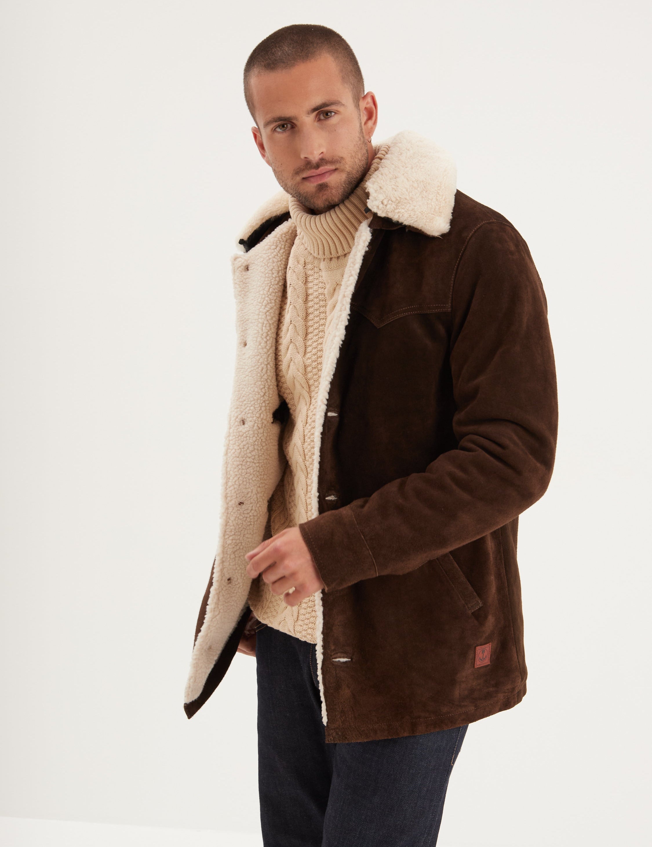 Man in a brown shearling coat and cream sweater stands against a white background, looking confidently at the camera.