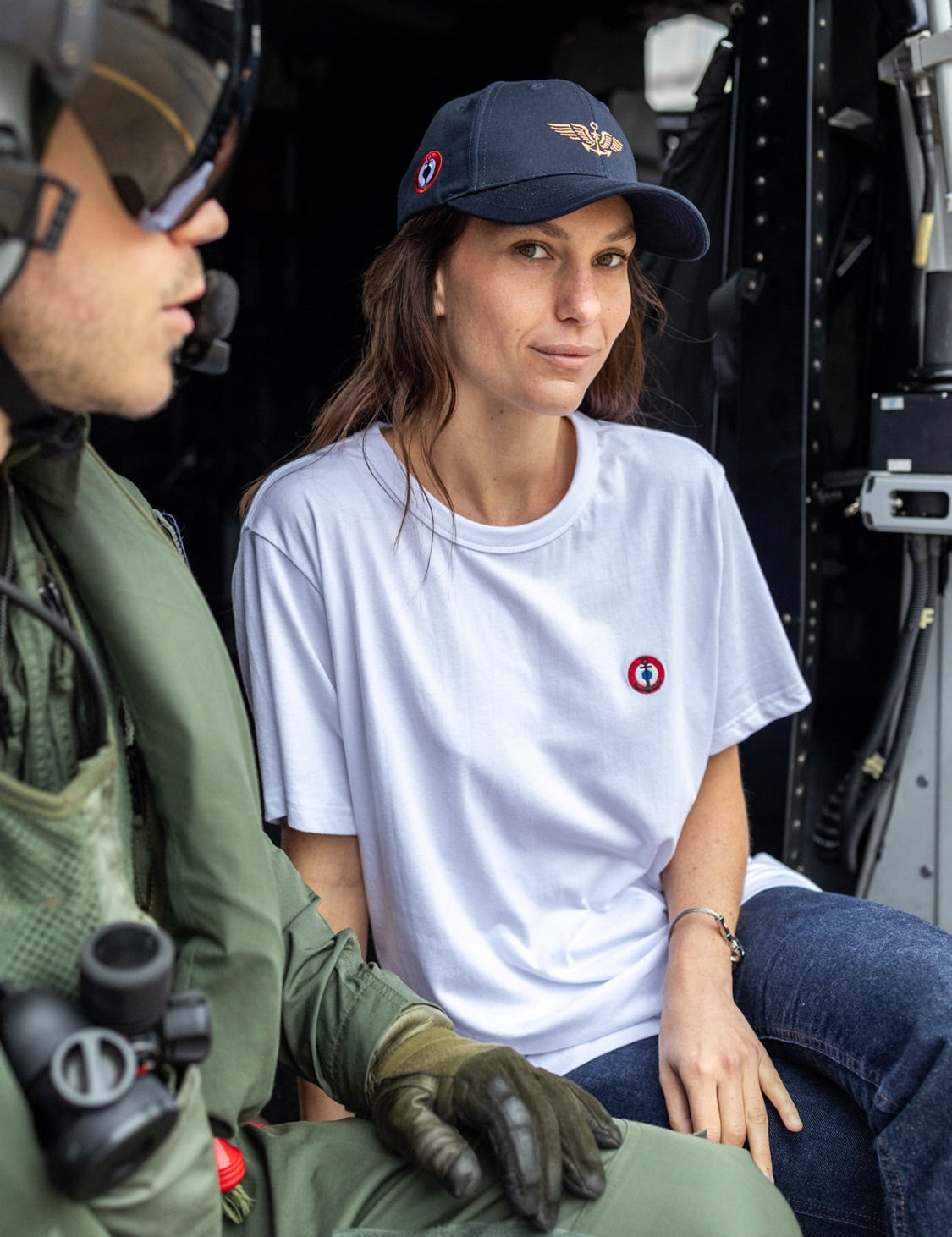 A woman in a white shirt and cap with an aviation logo sits beside a pilot in a helicopter, looking confidently at the camera.