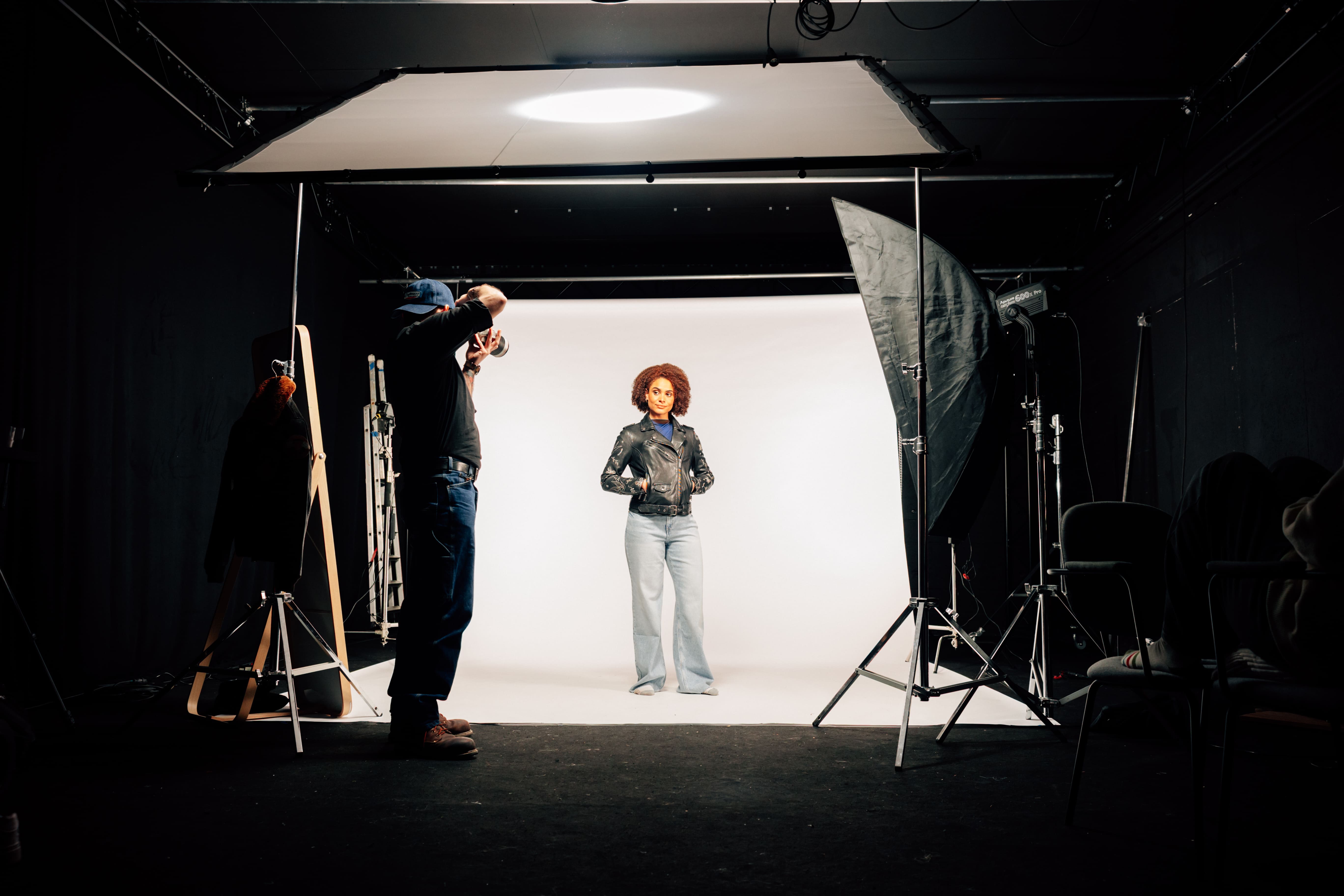 Photographer captures a model in a leather jacket and jeans under studio lights, set against a white backdrop in a dark studio.