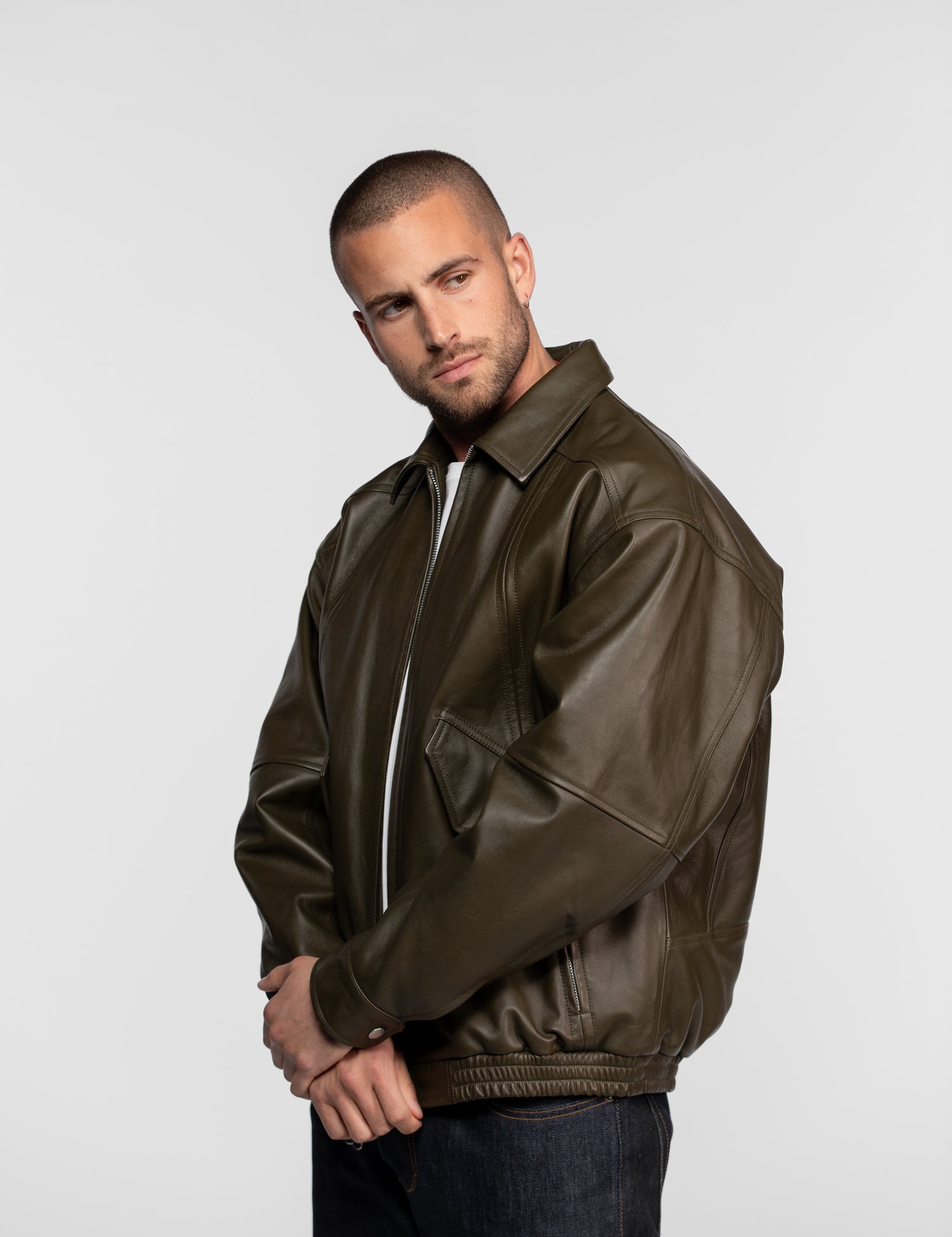 Man in a brown leather jacket stands confidently against a plain background, hands clasped, looking to the side.