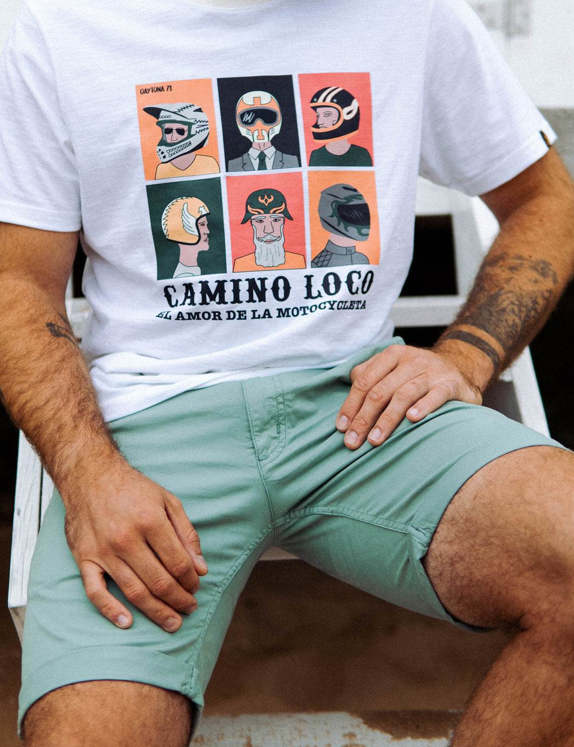 Man in green shorts sits wearing a white "Camino Loco" shirt with colorful helmet illustrations, celebrating motorcycle love.