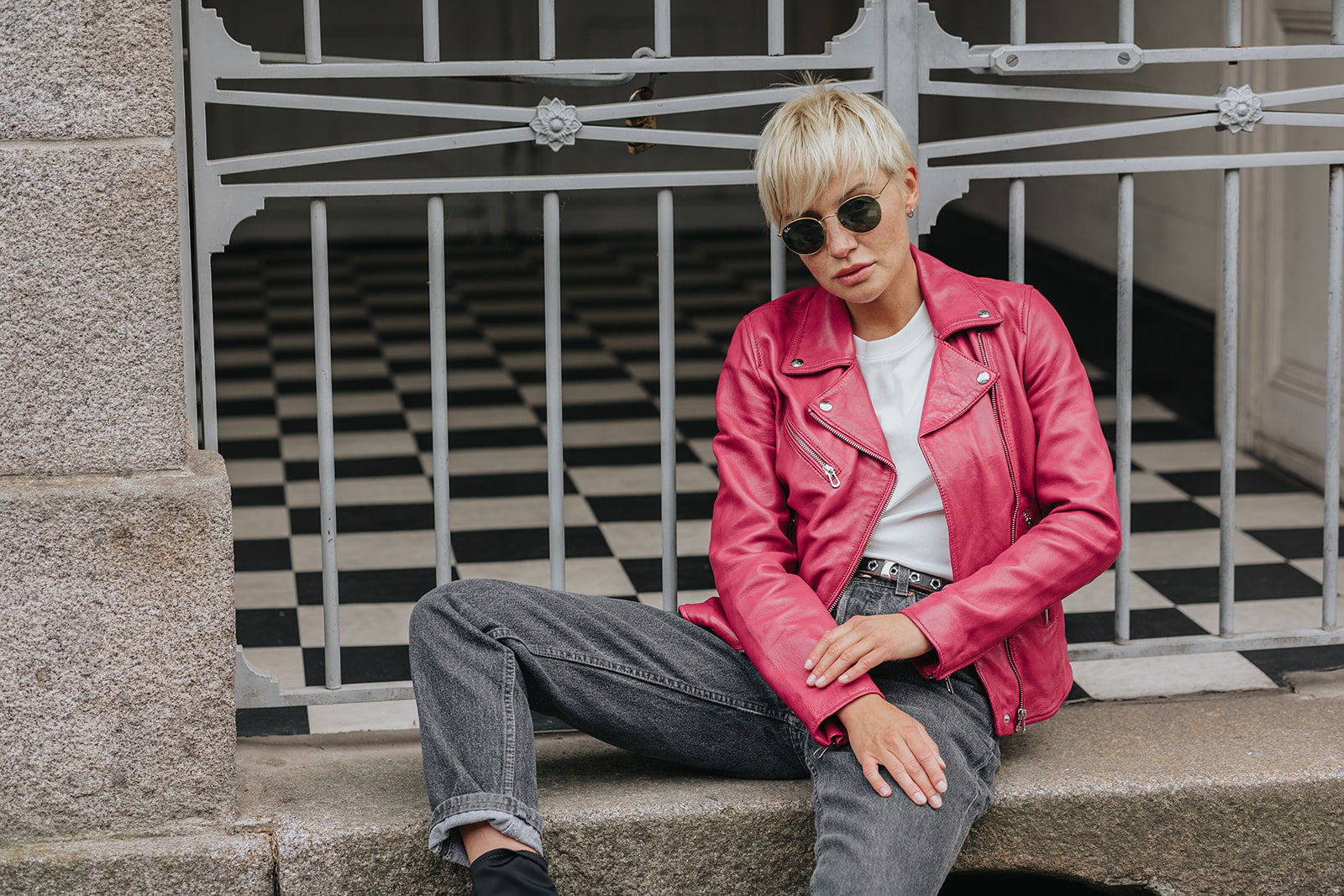 Person in pink jacket and sunglasses sits on a ledge, black and white checkered floor behind a metal gate in the background.
